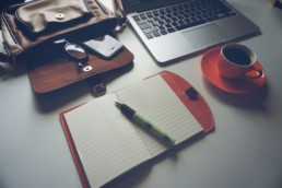 un tavolo con poggiati sopra, in maniera ordinata, un computer portatile una tazzina di caffè un piccolo quaderno ed uno zainetto posizionati in maniera ordinata