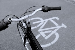 una bici della quale si vede la parte anteriore, quindi il manubrio, la ruota anteriore e parte del telaio. Sullo sfondo si vede l'asfalto con al di sopra di esso il disegno (segnaletica orizzontale) rappresentante una bicicletta stilizzata