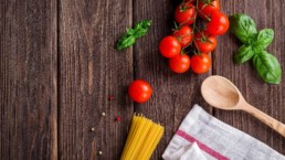 Tavolo di legno fotografato dall'alto. Su di esso sono presenti una mappina, degli spaghetti, dei pomodorini, del basilico ed un cucchiaio di legno