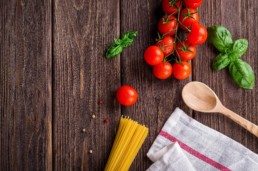 Tavolo di legno fotografato dall'alto. Su di esso sono presenti una mappina, degli spaghetti, dei pomodorini, del basilico ed un cucchiaio di legno